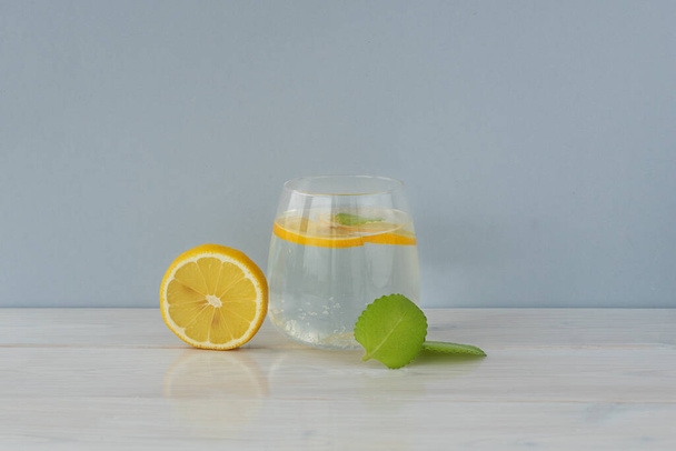 Прозрачный стакан воды, круглый ломтик лимона и мятный лист на светлом фоне. Детоксикационные витаминные напитки. - Фото, изображение
