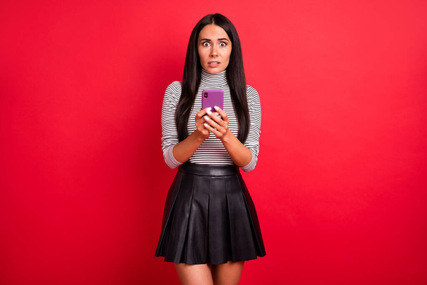 Portrait of attractive worried puzzled long-haired girl using device browsing fake news post isolated over bright red color background - Photo, Image