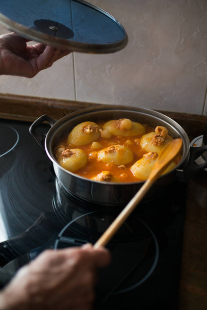 Típico plato asturiano. Manos de mujer envejecida con una olla y una cuchara de madera mientras se cocinan patatas llenas de atún, tomate y huevos. Gastronomía en casa. - Foto, imagen