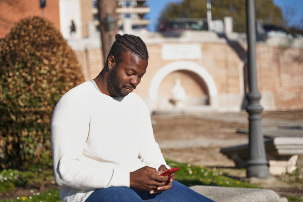 Junger Afroamerikaner sitzt auf einer Parkbank und sendet eine Botschaft aus. Latino-Mann mit Anti-Ansteckungsmaske. Konzept der neuen Normalität.  - Foto, Bild