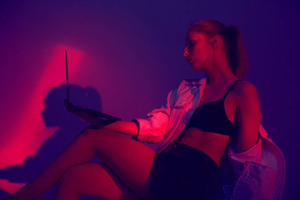 Young pretty girl create cool project on laptop in colourful neon light. Fashion model with computer - Foto, Imagem