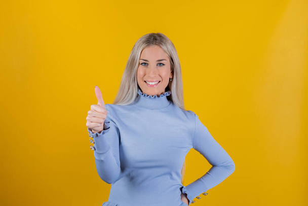 Woman satisfied with good quality of product, dressed in casual clothes, model against yellow background - Positive girl showing thumb up and looking at the camera - Photo, Image