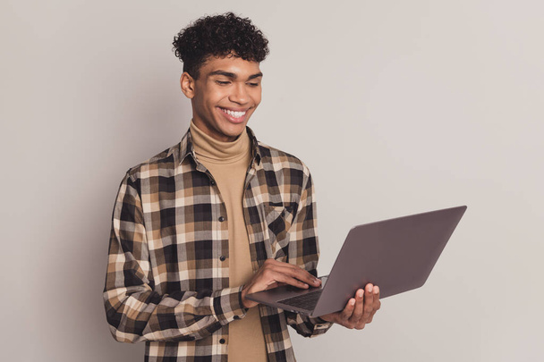 Foto retrato de un niño joven con camisa a cuadros trabajando en el ordenador portátil haciendo proyecto sonriendo aislado sobre fondo de color gris - Foto, Imagen
