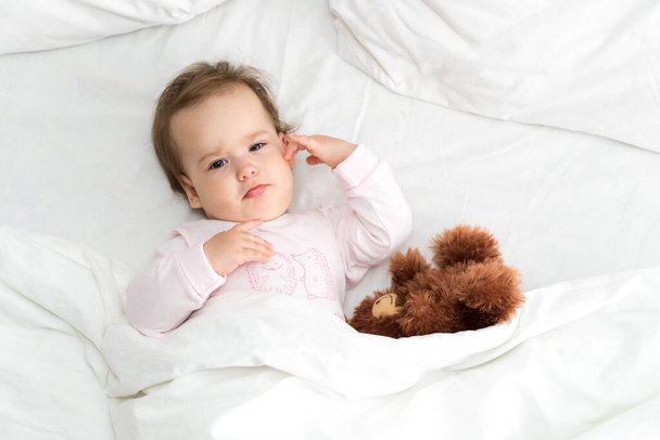Hiteles portré aranyos kaukázusi kis csecsemő pufók kislány vagy fiú rózsaszín álmos ébredés után maci nézi kamera fehér ágyban. Gyermekgondozás, Gyermekkor, Szülőkor, életmód koncepció - Fotó, kép