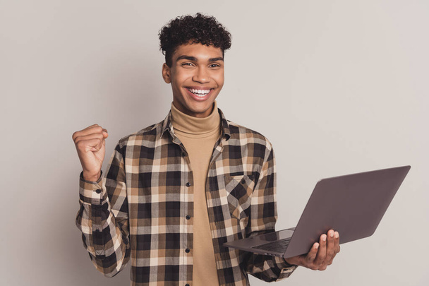 Photo of cheerful guy raise fist hold laptop beaming smile wear plaid shirt turtleneck isolated grey color background - Photo, image