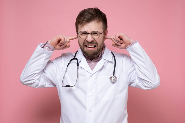 Medico con uno stetoscopio intorno al collo è stressato, è infastidito dal rumore, copre le orecchie con dita e denti sporgenti.  - Foto, immagini