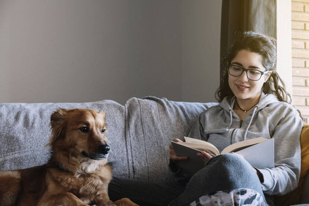 Молода жінка читає книгу, що сидить на дивані зі своїм собакою. Розслабтеся концепції
. - Фото, зображення