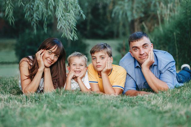 Όμορφη ευτυχισμένη Καυκάσια οικογένεια τεσσάρων ατόμων ξαπλωμένη σε ένα έδαφος στο πάρκο την καλοκαιρινή μέρα. Μητέρα και πατέρας με δύο γιους αδέλφια αδέλφια στην ύπαιθρο. Lifestyle αυθεντικά φυσικά συναισθήματα στιγμή.  - Φωτογραφία, εικόνα