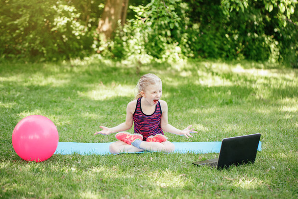Angry mad girl child doing sport workout outdoors online. Video yoga class on Internet. Funny stressed kid training in park with laptop. New normal. Poor Internet connection - Photo, Image