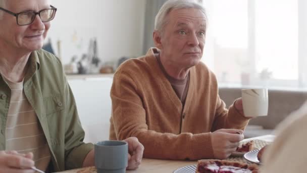 Inclinazione di due anziani caucasici seduti a tavola a casa, mangiare dolci, bere tè, parlare con persone irriconoscibili - Filmati, video