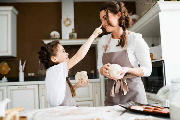 A sweet daughter touches her finger in flour to the nose of her mother who sculpts cookies from the dough - Foto, Imagem