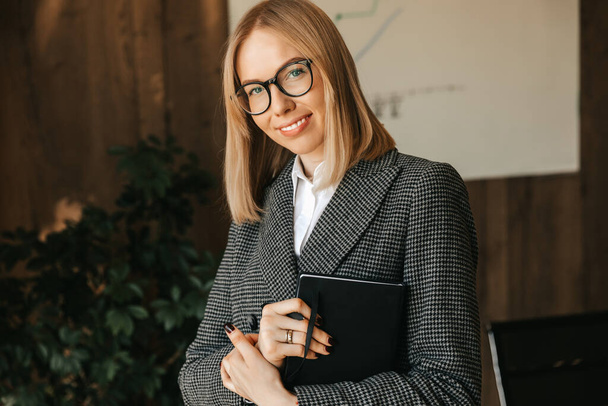 Μια επιχειρηματίας κρατά ένα σημειωματάριο στα χέρια της στέκεται και χαμογελά στο γραφείο ενώ σχεδιάζει νέα έργα για την εταιρεία της. Υψηλής ποιότητας φωτογραφία - Φωτογραφία, εικόνα