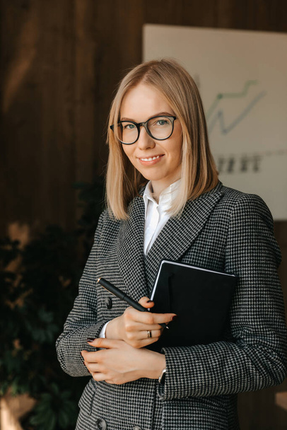 Μια επιχειρηματίας κρατά ένα σημειωματάριο στα χέρια της στέκεται και χαμογελά στο γραφείο ενώ σχεδιάζει νέα έργα για την εταιρεία της. Υψηλής ποιότητας φωτογραφία - Φωτογραφία, εικόνα