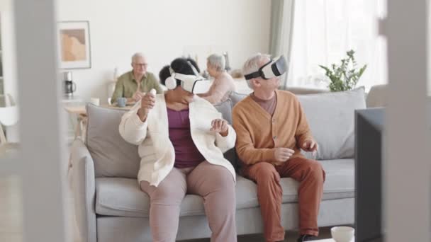 Medium lang van geamuseerde man en vrouw dragen virtual reality headsets, zitten op de bank in de woonkamer, bewegende hoofden rond, glimlachen - Video
