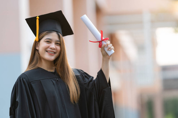 Młoda, szczęśliwa Azjatka, absolwentka uniwersytetu w sukni dyplomowej i moździerzowej posiada dyplom z wyróżnieniem za osiągnięcia edukacyjne w kampusie uniwersyteckim. Zdjęcie edukacyjne - Zdjęcie, obraz