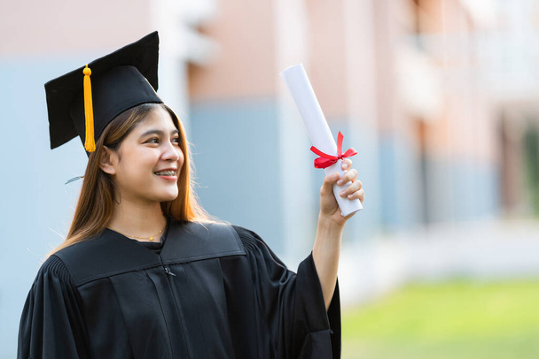 Mezuniyet elbisesi ve havan tahtasıyla genç mutlu bir Asyalı kadın üniversite mezunu üniversite kampüsünde eğitim başarısını kutlamak için diploma sahibi. Eğitim stoku fotoğrafı - Fotoğraf, Görsel