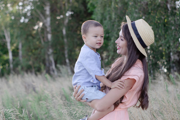 happy family - mother and son hug in the field. pretty brunette woman in cream dress and straw hat and little boy in blue costume. parenting, motherhood - Zdjęcie, obraz