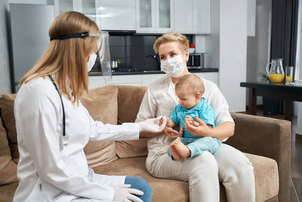 Competent doctor in mask examining baby at home - Foto, afbeelding