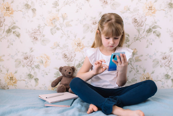 小さな女の子は、スタイリッシュなベッドルームのベッドの上に座って電話を持ち、スマートフォンで何かを読み取ります。コミュニケーションの概念 - 写真・画像