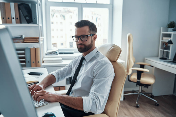 Молодий розумний чоловік у формальному одязі, що працює на комп'ютері, сидячи в офісі
 - Фото, зображення