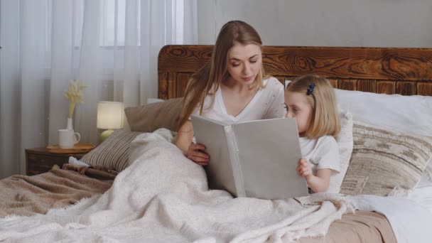 Кавказская молодая взрослая мать-одиночка мама читает сказочную книгу возлюбленной дочурке девочке ребенку-малышу ночью перед сном семья лежит в постели в уютной спальне на дому вечером - Кадры, видео