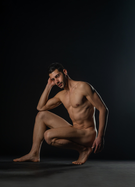 Retrato de una modelo masculina desnuda posando en cuclillas sobre fondo negro del estudio - Foto, imagen