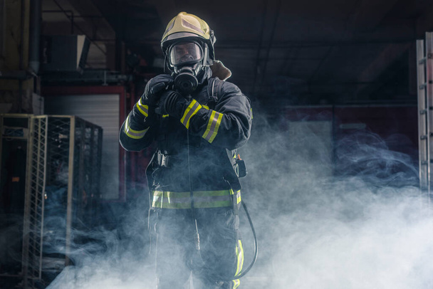 Portré egy tűzoltóról, aki tűzoltó felszerelést és sisakot visel. Sötét háttér füst és kék fény. - Fotó, kép