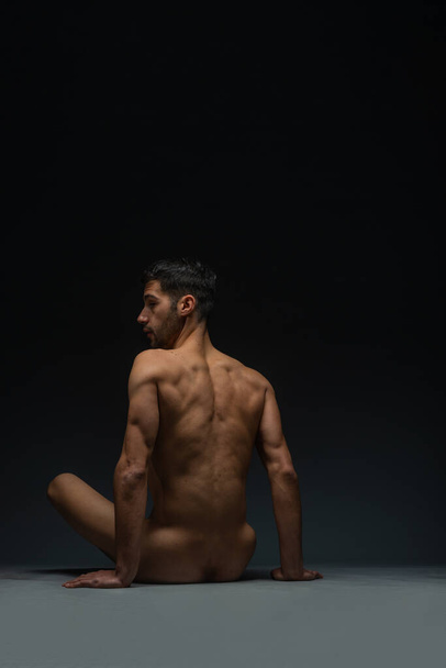 Вид ззаду гладкий голий спокусливий чоловік, що сидить на підлозі в студії, ізольований на чорному
 - Фото, зображення