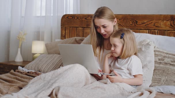 Young adult woman mom Caucasian mother hugs cuddles little daughter child baby look together in laptop screen choose movie online shopping in net make video call talking smiling lying in family bed - Footage, Video