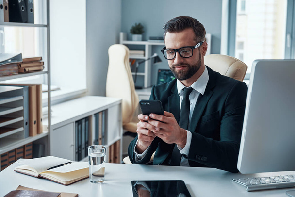 Good looking young man in shirt and tie using smart phone and smiling while sitting in the office - Fotoğraf, Görsel