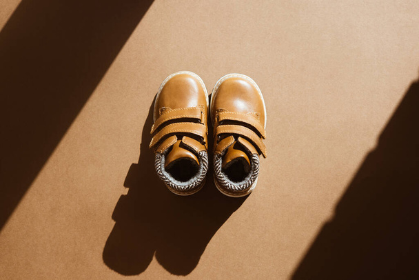 A pair of small leather shoe on a light, brown background with sun shadow. Warm, fur-lined children's shoes with laces. Concept of the coming autumn and winter top view - Zdjęcie, obraz