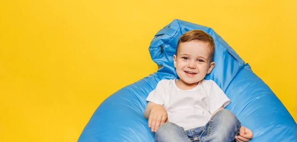 Lachender kleiner Junge auf gelbem Hintergrund, der in einem blauen Stuhl sitzt. Kopierraum. - Foto, Bild