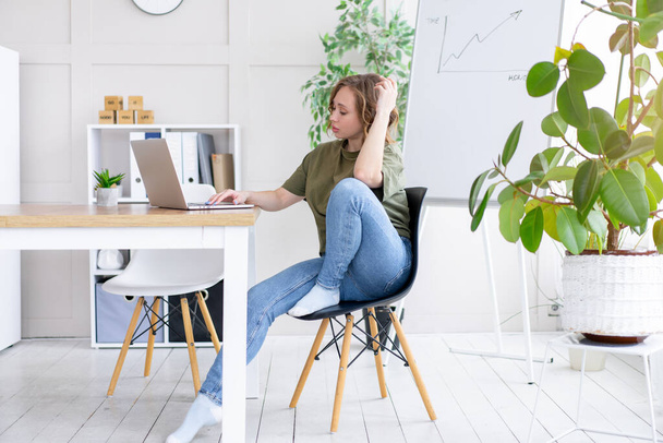 Business woman using laptop sitting desk white office interior with houseplant looking Business people Business person Online, Young and successful Dresed green shirt blue jeans barefoot relaxing  - Fotó, kép