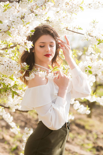 Lovely young girl in a white romantic silk blouse in spring in a blooming cherry garden in sunny weather. Spring and Fashion - Foto, afbeelding