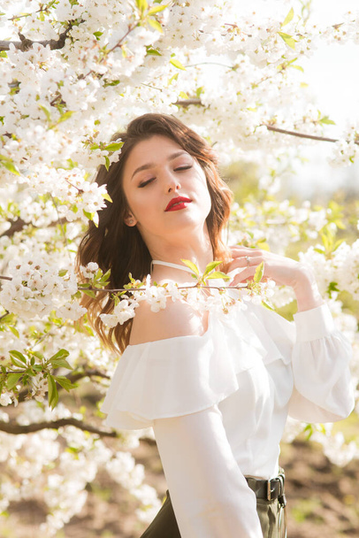 Piękna młoda dziewczyna w białej, romantycznej jedwabnej bluzce wiosną w kwitnącym wiśniowym ogrodzie w słoneczną pogodę. Wiosna i moda - Zdjęcie, obraz