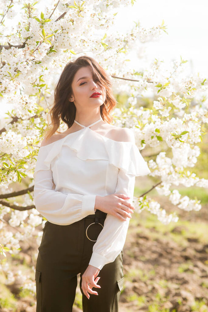 Lovely young girl in a white romantic silk blouse in spring in a blooming cherry garden in sunny weather. Spring and Fashion - Foto, imagen