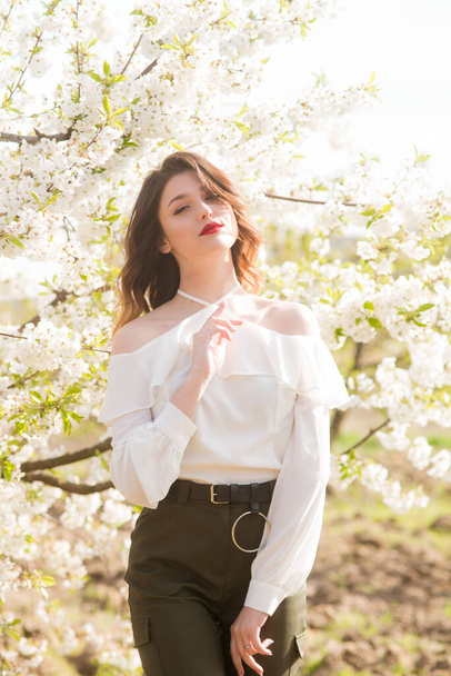 Lovely young girl in a white romantic silk blouse in spring in a blooming cherry garden in sunny weather. Spring and Fashion - Valokuva, kuva
