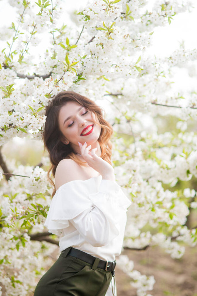 Lovely young girl in a white romantic silk blouse in spring in a blooming cherry garden in sunny weather. Spring and Fashion - Foto, Imagen