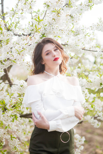 Lovely young girl in a white romantic silk blouse in spring in a blooming cherry garden in sunny weather. Spring and Fashion - Φωτογραφία, εικόνα