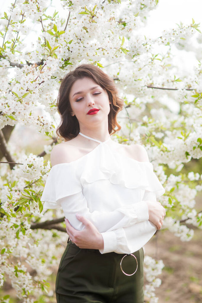 Lovely young girl in a white romantic silk blouse in spring in a blooming cherry garden in sunny weather. Spring and Fashion - Photo, image