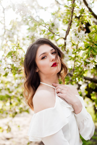 Lovely young girl in a white romantic silk blouse in spring in a blooming cherry garden in sunny weather. Spring and Fashion - Fotografie, Obrázek
