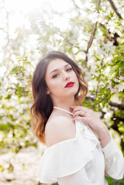 Gyönyörű fiatal lány egy fehér romantikus selyem blúzban tavasszal egy virágzó cseresznyekertben napsütéses időben. Tavasz és divat - Fotó, kép