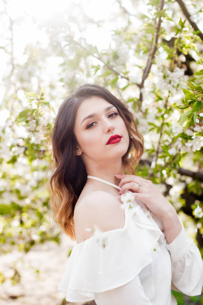 Lovely young girl in a white romantic silk blouse in spring in a blooming cherry garden in sunny weather. Spring and Fashion - 写真・画像