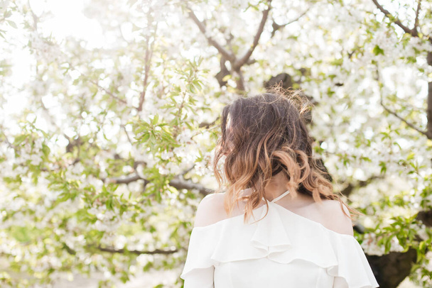 Υπέροχο νεαρό κορίτσι σε ένα λευκό ρομαντικό μεταξωτό πουκάμισο την άνοιξη σε ένα ανθισμένο κήπο κεράσι σε ηλιόλουστο καιρό. Άνοιξη και μόδα - Φωτογραφία, εικόνα