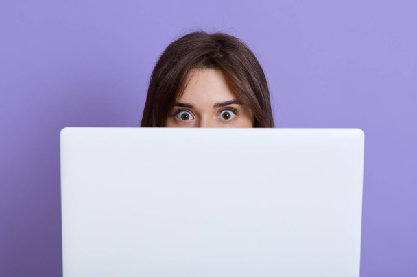 Scared dark haired female hiding behind laptop and looking at camera with eyes full of fear, being afraids of something, wants somebody do not see her, isolated over lilac background. - Фото, изображение