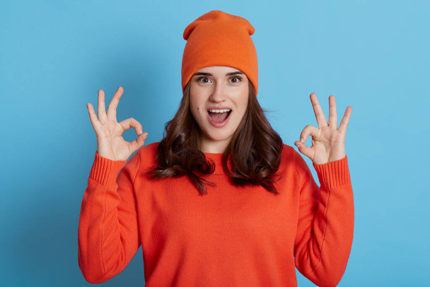 Young European excited female wearing orange sweater and cap looking at camera with opened mouth and showing ok signs with both hands, dark haired girl isolated over blue background. - Φωτογραφία, εικόνα