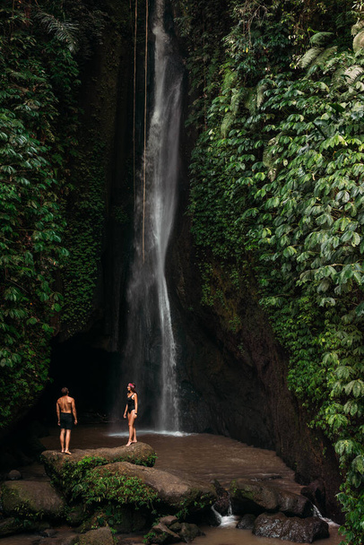 Couple at a tropical waterfall. The couple travels to the island of Bali in Indonesia. A woman and a man at a beautiful waterfall. Vacation on a tropical island. Traveling couple at the waterfall - Fotografie, Obrázek