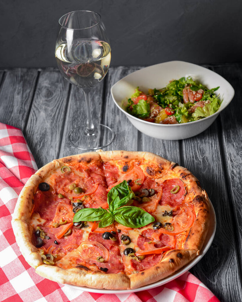 Fresh italian pizza with salami and black olives served with a glass of white wine. Traditional Italian pizza recipe, Italian cuisine. Close up on pizza. Fast food, junk food concept. - Фото, зображення