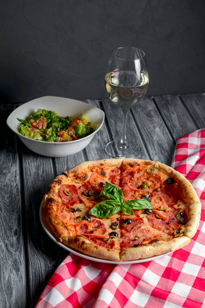 Fresh italian pizza with salami and black olives served with a glass of white wine. Traditional Italian pizza recipe, Italian cuisine. Close up on pizza. Fast food, junk food concept. - Photo, Image