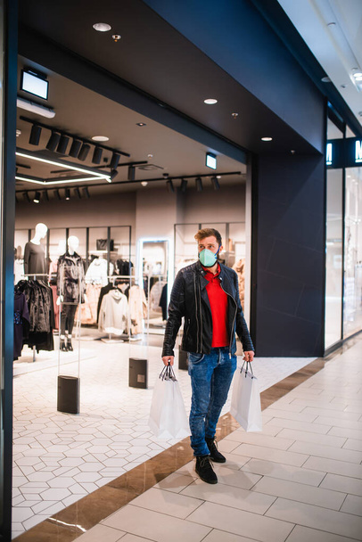 Um jovem caucasiano elegante em uma jaqueta de couro, com uma máscara e bolsas na mão saindo de uma boutique em um shopping durante uma epidemia do coronavírus COVID-19 - Foto, Imagem